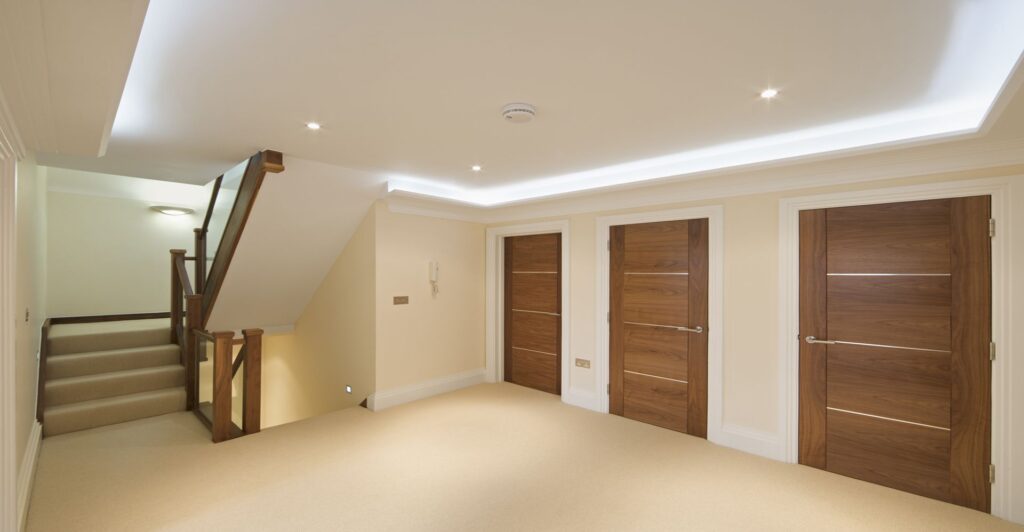 Un corridoio con pareti e moquette color crema, tre porte artigianali chiuse in legno sulla destra e una scala con ringhiera in legno sulla sinistra. I lampadari a soffitto forniscono illuminazione.